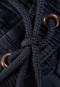 Tela negra con textura tejida, que presenta una cuerda negra torcida y un ojal de bronce. El nudo es prominente y está bien definido.