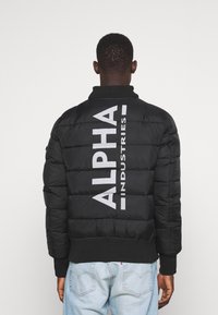 Mand iført en sort polstret jakke med stor hvid "ALPHA INDUSTRIES" tekst på ryggen, parret med lyseblå jeans.