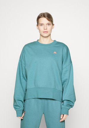 Mėlynos spalvos per didelis džemperis su apvaliu kaklu, nuleistomis petnešomis ir mažuoju koralinės spalvos logotipu kairėje krūtinės pusėje. Lygus audinio paviršius.