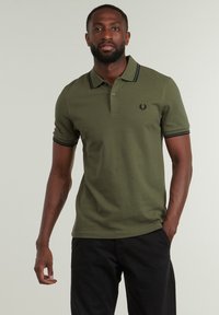 Camiseta polo verde oliva con ribetes negros en el cuello y las mangas, que presenta un pequeño logo bordado en el pecho, hecha de una tela transpirable.
