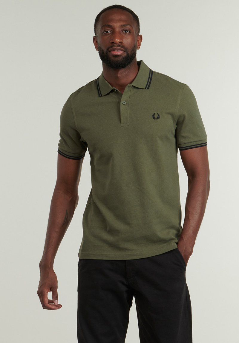 Camiseta polo verde oliva con ribetes negros en el cuello y las mangas, que presenta un pequeño logo bordado en el pecho, hecha de una tela transpirable.
