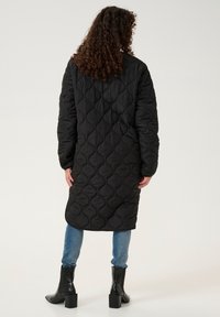 Černý prošívaný dlouhý kabát s texturovaným vzorem, volného střihu, nošený přes světlé modré džíny a černé kotníkové boty.