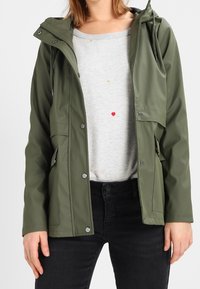 Grøn vandtæt jakke med hætte, der har trykknapper og sidelommer. Båret over en lysegrå sweater med små farvede detaljer.