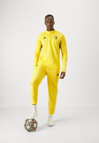 Chándal deportivo amarillo con accents negros, cuello con cremallera y tres rayas en las mangas. De pie sobre un balón de fútbol con zapatillas blancas.