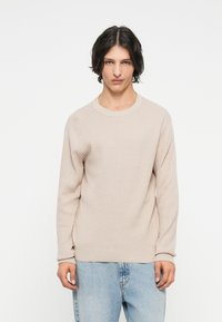 Beige kötött pulóver bordázott textúrával, kerek nyakkivágással és hosszú ujjúakkal, világoskék farmerrel párosítva. Minimalista dizájn.