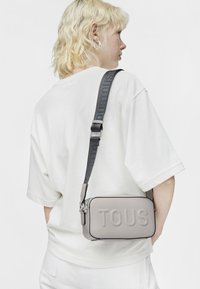 Borsa a tracolla quadrata in materiale grigio chiaro testurizzato con logo "TOUS" impresso, caratterizzata da una tracolla scura a contrasto con brand.