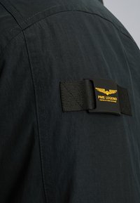 Zwarte stoffen kledingstuk met zichtbare stiksels, voorzien van een zwarte rechthoekige label met gele tekst en een vleugelgrafiek die prominent is bevestigd.
