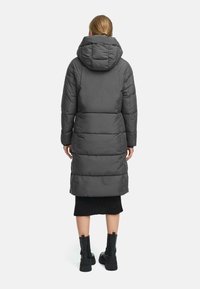 Cappotto puffer grigio con cappuccio, caratterizzato da un design trapuntato, lunghezza fino al ginocchio e dettagli di cucitura in evidenza. Indossato sopra una gonna nera a coste.