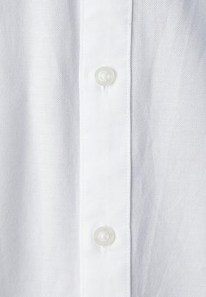 Baltas marškinys iš tekstūruotos medžiagos, su tiesiu plokšteliu priekyje, kurioje yra dvi skaidrios, apvalios sagos.