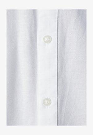 Baltas marškinys iš tekstūruotos medžiagos, su tiesiu plokšteliu priekyje, kurioje yra dvi skaidrios, apvalios sagos.