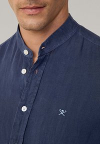 Camicia blu navy con colletto alla cinese, bottoni bianchi e piccolo dettaglio ricamato di oggetti incrociati sul petto.