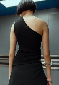 Person mit kurzem schwarzen Haar in einem schwarzen Top mit einem Schulterträger und schwarzen Hosen, steht drinnen mit grauen Paneelwänden und einer Deckenleuchte darüber.