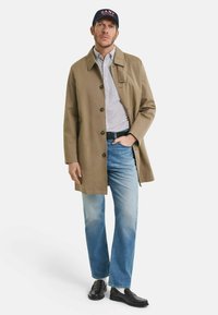 Man in een beige jas, gestreept shirt, blauwe jeans, zwarte loafers, witte sokken en een marineblauwe pet met een rood logo, staande met één hand in zijn zak.