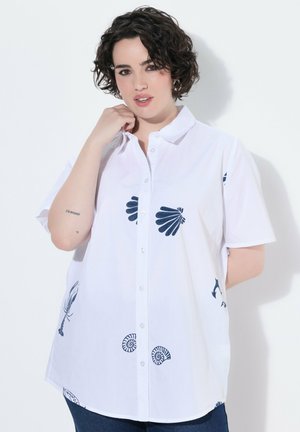 Jonge vrouw met kort krullend donker haar, gekleed in een wit blouse met marineblauwe schelp- en zeemotieven, poserend met één hand bij haar gezicht.
