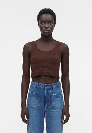 Άτομο με σκούρο τόνο δέρματος που φοράει αμάνικη καφέ κοντή μπλούζα και τζιν ψηλόμεσο, στέκεται μπροστά σε απλό φωτεινό φόντο.