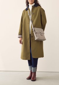 Olivevärvi mantel tumerohelise kraega, hall käekott, kitsas valge kaelusega sviiter ja tumedad teksad siniste mansettidega; pruunid pahkluu saapad.