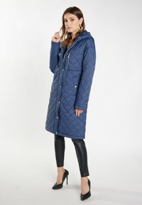 Cappotto lungo trapuntato blu navy con cappuccio, dotato di tasche con zip, chiusura a bottoni e tessuto texture. Abbinato a pantaloni neri aderenti.