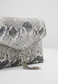 Clutch-laukku harmaassa ja mustassa käärmekuvioidussa nahassa, jossa on taitettava läppä ja hopeinen hapsukoriste kiinnitetty suljettavaan osaan. Pinnan tekstuuri on elävä.