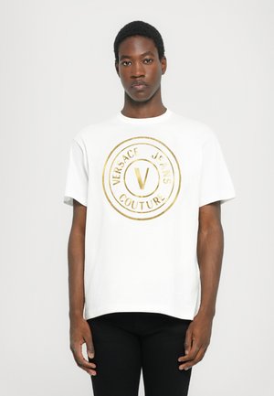 Biała bawełniana koszulka z dużym złotym okrągłym logo z napisem „VERSACE JEANS COUTURE” oraz widoczną literą „V” na środku.
