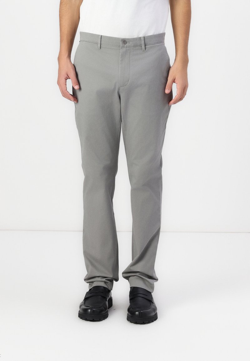 Homme portant un pantalon droit gris clair, des chaussures noires et une chemise blanche, debout devant un fond blanc uni.