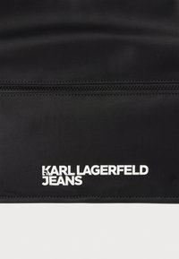 Μαύρο νάιλον ύφασμα με φερμουάρ και κείμενο "KARL LAGERFELD JEANS" με έντονα λευκά γράμματα. Το κείμενο βρίσκεται κοντά στο φερμουάρ.