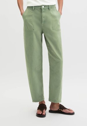 Personne portant un pantalon droit vert clair avec des poches plaquées à l'avant et des sandales marron foncé, debout les mains dans les poches.