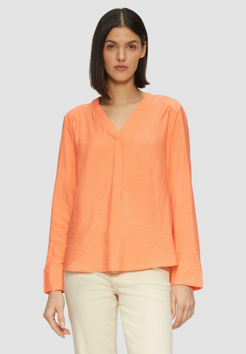 T-shirt à manches longues en V de couleur corail vif, fabriqué dans un tissu texturé léger avec une coupe décontractée et un détail de plis subtil.