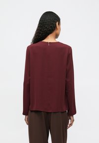 Γυναίκα με σγουρά μαλλιά, φορώντας μπλούζα σε μπορντό απόχρωση με μακρύ μανίκι και καφέ παντελόνι, προφίλ από πίσω σε απλό φόντο.