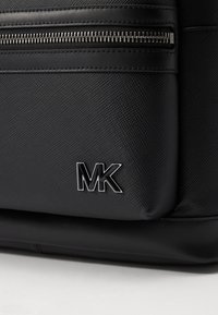 Černá syntetická kabelka s texturovaným povrchem, zipovým uzávěrem a stříbrnými detaily. Logo "MK" výrazně umístěné na boku.