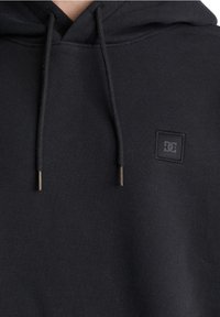 Sweat à capuche noir avec une poche kangourou à l'avant, des cordons ajustables et un petit patch logo noir sur la poitrine. Tissu doux et texturé.