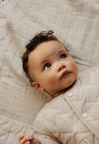 Veste pour bébé matelassée beige avec boutons-pression, présentant une texture douce et un col arrondi, posée sur une couverture texturée neutre. Bébé regarde vers le haut.