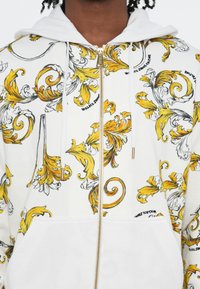 White zip-up hoodie with yellow and black baroque pattern, featuring a front pocket, drawstrings, and gold hardware accents.