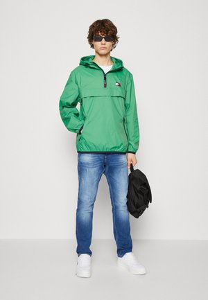 Chaqueta con capucha ligera y verde con un cierre de cuarto de cremallera en la parte frontal, que presenta un parche con el logo. Combinada con unos vaqueros azules y zapatillas blancas.