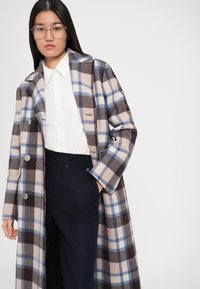 Manteau à carreaux aux tons beige, marron et bleu, fabriqué à partir d'un tissu doux. Double boutonnage avec de grands boutons et un col cranté.
