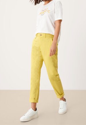 Femme portant un pantalon jaune retroussé, un t-shirt graphique blanc et des baskets blanches, se tenant devant un fond neutre.