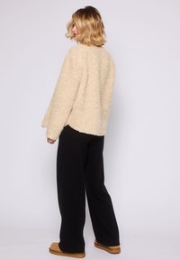 Donna in piedi di lato che indossa un maglione beige peloso, pantaloni neri a gamba larga e scarpe slip-on color beige su uno sfondo chiaro e semplice.
