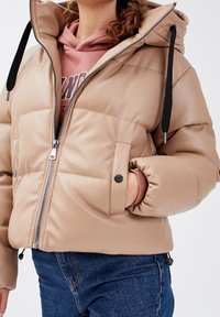 Doudoune beige à finition brillante, fermeture éclair, capuche avec cordons de serrage et coutures décoratives. Un jean bleu visible en dessous.