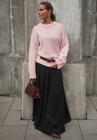 Sieviete rozā džemperī un melnā maxi svārkā, turot bordo rokassomu, stāv pie teksturēta betona sienas.