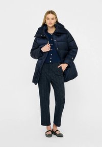 Tmavomodrý vatovaný kabát s vysokým golierom a horizontálnym prešitím. Kombinovaný s tmavomodrým pleteným topom a vzorovanými nohavicami. Čierne balerínky.