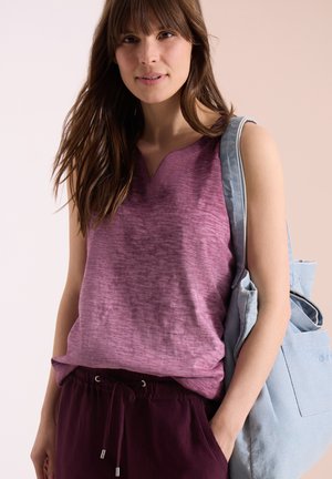 Jeune femme portant un haut violet sans manches et un pantalon foncé, portant un sac en toile bleu clair sur l'épaule.