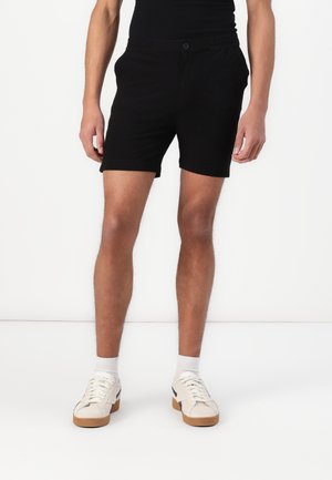 Shorts noirs en tissu texturé, avec une taille élastique et une fermeture à bouton. Portés avec des baskets blanches et des chaussettes.