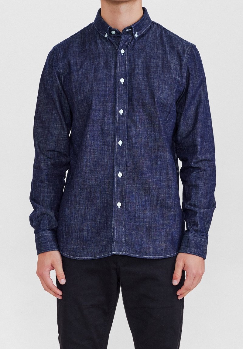 Chemise en denim foncé avec un col boutonné, des manches longues et des boutons blancs. Caractérisée par un tissu texturé et des détails de couture visibles.