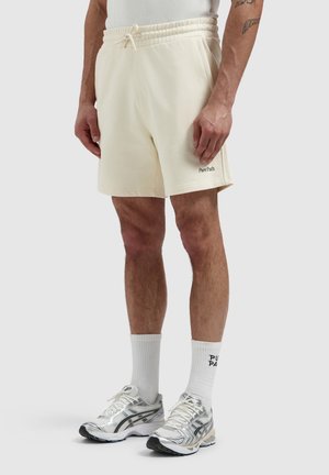 Man draagt crèmekleurige short met trekkoord en "Pure Path" logo, witte "Pure Path" sportsokken en zilveren hardloopschoenen, staand.