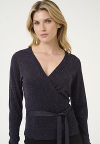Pull enroulé violet foncé avec manches longues, col en V, tissu texturé et taille nouée. Finition légère pailletée créant un effet scintillant.
