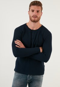 Mornarsko modra dolgosleeve henley majica z teksturirano tkanino, okroglo izrezano ovratnikom, tri gumbi na prednji strani in zoženimi rokavi, v kombinaciji z džinsom.