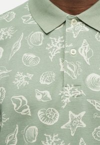 Πράσινο πόλο μπλουζάκι με λευκά σχέδια κοχυλιών και κοραλλιών, με γιακά και δύο λευκά κουμπιά κοντά στον λαιμό.