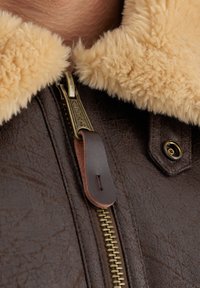 La chaqueta de cuero marrón cuenta con un cuello de piel sintética beige, un zipper de latón con una lengüeta de cuero, y un detalle de botón a presión.