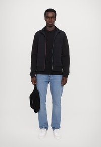 Svart quiltet vest over en svart hettegenser, parret med lyseblå jeans og hvite sneakers. Holder en svart skulderveske.