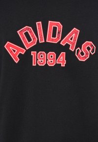 Μαύρο βαμβακερό μπλουζάκι με μεγάλο κόκκινο κείμενο που γράφει «ADIDAS» κυρτό πάνω από το «1994», με μια φθαρμένη υφή στα γράμματα.