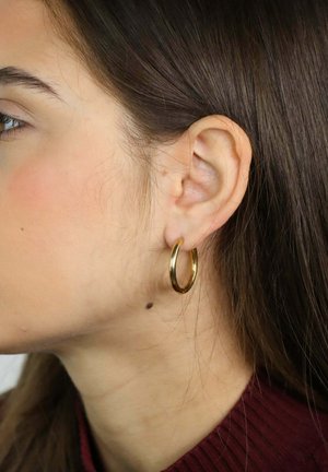 Jeune femme aux longs cheveux bruns portant une boucle d'oreille en or, oreille gauche et cou visibles, vêtue d'un haut bordeaux.